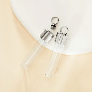 Peut inclure: Deux flacons en verre argentés avec des bouchons métalliques et une boucle pour accrocher. Les flacons sont transparents et vides.
