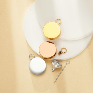 May include: Three round, metallic pendants in gold, rose gold, and silver, each with a small loop for a chain. A small silver funnel and a thin silver tool are also present, suggesting a jewelry or keepsake theme.