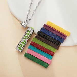 Può includere: Una collana diffusore di oli essenziali argentata con un design floreale, aperta per rivelare un interno verde. Una collezione di strisce di feltro colorate in varie tonalità, tra cui verde, rosa e giallo, sono disposte nelle vicinanze. La collana è su una superficie bianca.