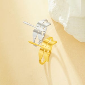 May include: Two dragonfly rings, one silver and one gold, are shown against a soft, neutral background. The rings feature a delicate dragonfly design, with wings and body details. The gold ring has a shiny finish, while the silver ring has a matte appearance.