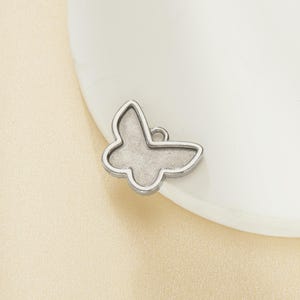 Könnte beinhalten: Ein kleiner, silberfarbener Schmetterlingsanhänger mit einer Schlaufe zum Befestigen an Schmuck. Der Anhänger hat eine detaillierte Kontur und eine glatte, matte Oberfläche. Der Schmetterling liegt auf einer weißen Oberfläche.