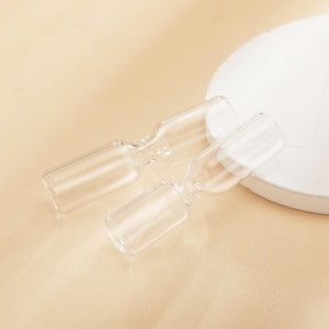 Puede incluir: Dos viales de vidrio transparente con una abertura acampanada y un cuello estrecho. Los viales están sobre una superficie beige claro junto a un objeto blanco y redondo. Los viales están vacíos y parecen ser para uso de laboratorio o cosmético.