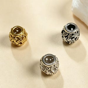 May include: Three spherical beads with an intricate openwork floral pattern. One bead is gold-toned, one is dark gunmetal grey, and one is bright silver-toned. Each bead features a smooth, circular opening at the top and bottom for threading.