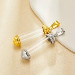 May include: Two glass vial pendants with metal caps and loops. One has a gold-tone cap and loop, the other a silver-tone cap and loop. The cylindrical vials are empty, ideal for small keepsakes or charms. The pendants are displayed on a cream-colored surface.