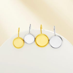 May include: Four round, blank, metal earring settings. Two are gold and two are silver. The settings are on a white surface.