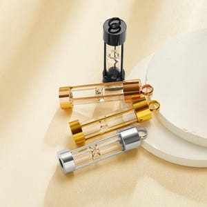 May include: An assortment of small hourglass pendants in gold, silver, and black. The pendants have clear glass tubes with a star-shaped design inside. The hourglasses are displayed on a neutral background.