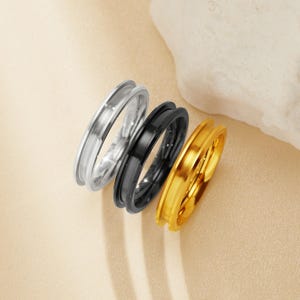May include: Three simple wedding bands in silver, black, and gold. The bands are stacked on top of each other, with the gold band on top.