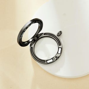 Könnte beinhalten: Ein schwarzes Metall-Medaillon mit einem Scharnierdeckel und einem transparenten Glaseinsatz. Das Medaillon ist geöffnet und der Deckel liegt auf der Oberfläche.