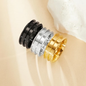 May include: Three stacked rings, one black, one silver, and one gold. Each ring has a grooved design.