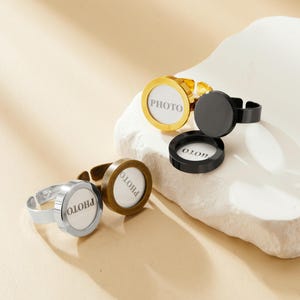 Könnte beinhalten: Vier verstellbare Ringe mit einem runden Bilderrahmen. Die Ringe sind golden, silber und schwarz. Die Bilderrahmen haben den Schriftzug "PHOTO" aufgedruckt.
