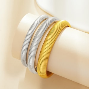 Könnte beinhalten: Drei Metallarmbänder in Silber und Gold. Die Armbänder haben ein geripptes, flexibles Design. Das goldene Armband befindet sich rechts, mit zwei silbernen Armbändern links. Die Armbänder werden auf einer cremefarbenen Oberfläche präsentiert.
