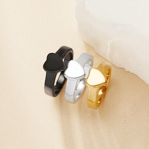 May include: Three heart-shaped rings stacked together. The top ring is black, the middle ring is silver, and the bottom ring is gold.