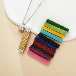 Op de afbeelding: Een zilverkleurige etherische olie diffuser ketting met een bloemenpatroon, open om een gele binnenkant te onthullen. Een verzameling kleurrijke vilten pads in roze, paars, rood, zwart, blauw, turquoise, geel en groen zijn in de buurt gerangschikt.