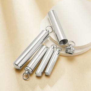 Puede incluir: Una colección de recipientes cilíndricos de metal plateado pulido con tapas de rosca y llaveros. Un recipiente está abierto, revelando el interior roscado. Los recipientes están dispuestos sobre una superficie blanca en capas.