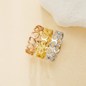 Może przedstawiać: Trzy złote pierścionki z wyciętym wzorem w kształcie serca. Pierścionki są ułożone jeden na drugim.