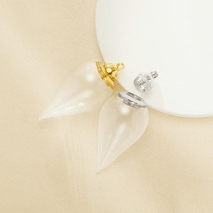 May include: Two clear glass teardrop-shaped pendants with gold and silver-coloured metal caps. One cap is gold, the other is silver. A small silver-coloured screw-on cap is also present. The pendants are on a cream-coloured surface.