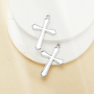 Puede incluir: Dos colgantes de cruz plateados con un centro blanco y un pequeño lazo en la parte superior para colgar. Las cruces se muestran sobre una superficie blanca en capas.