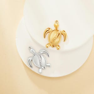 May include: Two turtle charms, one gold and one silver, with a detailed design on the shell. The charms are on a white surface.