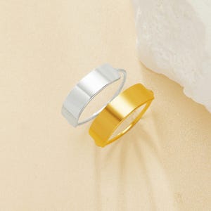 Può includere: Due anelli semplici a fascia piatta, uno dorato e uno argentato, impilati uno sopra l'altro. Gli anelli sono su uno sfondo beige chiaro.