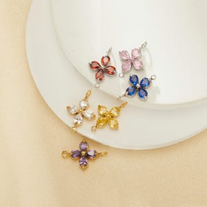 May include: A collection of flower-shaped jewelry charms in various colors, including red, pink, blue, white, yellow, and purple. Each charm features four gemstone petals and a small loop for attachment. The charms are displayed on a white surface.
