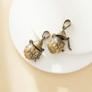 Puede incluir: Dos colgantes de metal dorado antiguo con diseños florales. Cada colgante es una pequeña caja redonda con bisagras con un patrón floral en el exterior. Las cajas están abiertas, revelando el interior vacío.