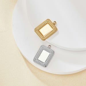 Puede incluir: Dos colgantes rectangulares, uno dorado y otro plateado, con un borde texturizado y un lazo para sujetar una cadena. Los colgantes se muestran sobre una superficie blanca.
