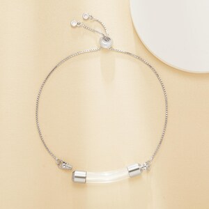 Puede incluir: Una pulsera plateada con una pieza central cilíndrica transparente. La pulsera presenta una delicada cadena y un cierre deslizante ajustable. El cilindro transparente tiene tapas plateadas. La pulsera se muestra sobre una superficie beige claro.