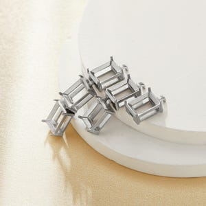 Puede incluir: Un grupo de engastes de piedras preciosas de metal plateado. Los engastes son de forma rectangular y tienen un acabado liso y pulido.