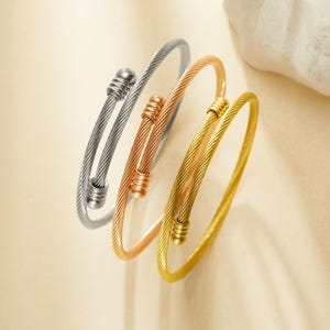 Könnte beinhalten: Drei Metallarmreifen in Silber, Roségold und Gold. Jedes Armband hat ein gedrehtes Kabeldesign und dekorative Endkappen. Die Armreifen werden auf einem neutralen Hintergrund präsentiert, der ihre verschiedenen metallischen Oberflächen hervorhebt.