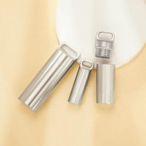 Puede incluir: Una colección de recipientes de acero inoxidable, que incluye una cápsula cilíndrica más grande con asa, una cápsula más pequeña y una tapa roscada. Los artículos están dispuestos sobre una superficie beige claro.