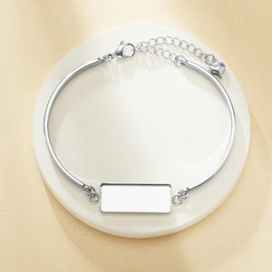 May include: A silver bracelet featuring a rectangular white centrepiece and a tiny heart charm. The bracelet has a curved design and a chain extension for adjustable sizing. The jewellery is displayed on a white surface.