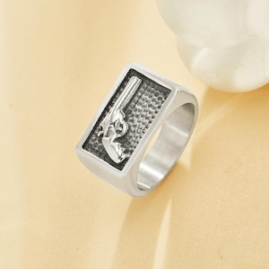 Puede incluir: Anillo plateado con una cara rectangular que presenta un diseño detallado de una pistola. El anillo tiene un acabado pulido y una banda gruesa. La pistola está representada en un estilo grabado detallado, sobre un fondo texturizado.