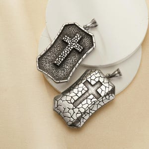 May include: Two silver-coloured pendant necklaces with cross designs. One pendant has a textured background and a cross with a dotted pattern. The other pendant has a cracked pattern background and a cross design.