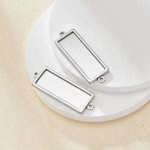 May include: Two silver-toned rectangular metal frames with open backs, each with two jump rings at the top. The frames are ideal for adding a personal touch to jewellery projects.