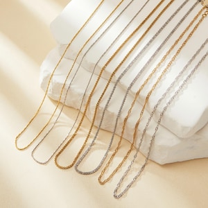 Può includere: Un set di sei collane a catena dorate e argentate di diverse larghezze e stili. Le catene sono disposte su una superficie bianca.