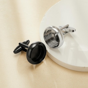 May include: Two cufflinks, one black and one silver, are displayed on a white surface. The black cufflink has a glossy finish, while the silver cufflink has a polished, metallic appearance. Both cufflinks have a circular design with a post for securing to a shirt cuff.