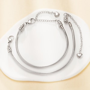 May include: Two silver bracelets with a snake chain design. One has a lobster clasp, the other an adjustable chain with a heart charm. The bracelets are displayed on a white plate. The chains are made of metal.