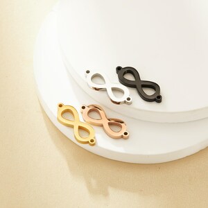 Könnte beinhalten: Drei Unendlichkeits-Anhänger aus Gold, Roségold und schwarzem Metall. Die Anhänger liegen auf einer weißen Oberfläche.