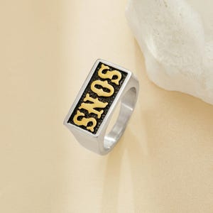 Peut inclure: Bague argentée avec une face rectangulaire portant l'inscription "SONS" en lettres dorées sur fond noir. La bague présente un design simple et moderne, adaptée à un usage quotidien.