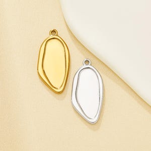 May include: Two irregular oval-shaped jewellery pendants, one in a polished gold tone and one in a polished silver tone. Each pendant features a smooth, recessed centre area surrounded by a raised border and a small circular loop at the top.