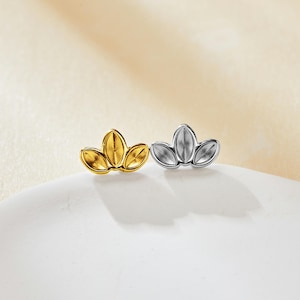 May include: Two stud earrings, one gold-toned and one silver-toned, each with a three-leaf design. The earrings are presented on a white surface against a soft, neutral background. The gold earring has a shiny finish.