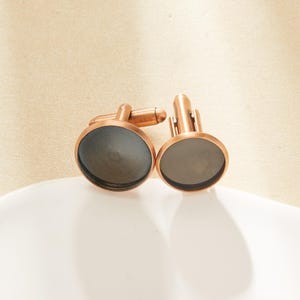 May include: A pair of copper-toned cufflinks with a dark grey, circular face. The cufflinks are presented on a white surface against a neutral background. The design features a classic, elegant style.
