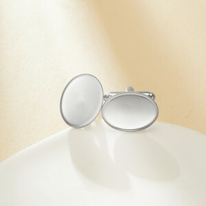 May include: A pair of silver-toned cufflinks with an oval, white face and a beaded edge. The cufflinks are displayed on a white surface against a soft, neutral background.