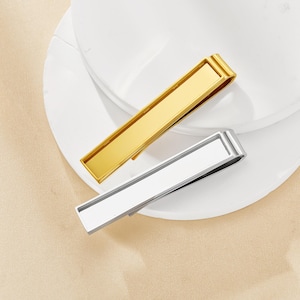 May include: Two metal tie clips, one gold and one silver, are displayed on a white surface. The gold clip has a shiny finish, while the silver clip has a matte appearance. Both clips are rectangular with a simple, modern design.