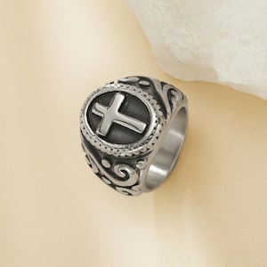 May include: A silver-toned ring with a prominent cross design. The ring features an oval centerpiece with a raised cross, surrounded by a beaded border and ornate scrollwork on the band. The ring is made of a shiny, metallic material.