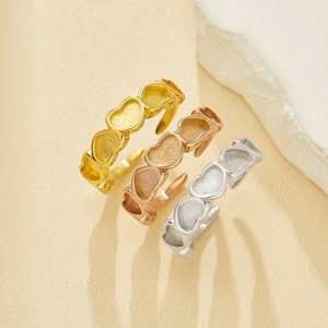Könnte beinhalten: Drei herzförmige Ringe in Gold, Roségold und Silber. Jeder Ring hat eine Reihe von verbundenen Herzen, die ein durchgehendes Design bilden. Die Ringe werden auf einer hellbeigen Oberfläche präsentiert.
