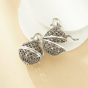 May include: Two silver-colored, filigree-style pendants. One is open, revealing the interior. The pendants have a hinged design and a small loop at the top for hanging. The intricate metalwork features a cross and rope pattern.