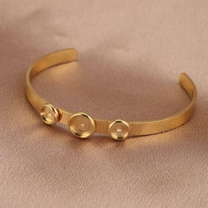 Può includere: Un braccialetto rigido dorato con tre accenti rotondi decorativi. Il braccialetto è aperto e ha un design semplice e minimalista.