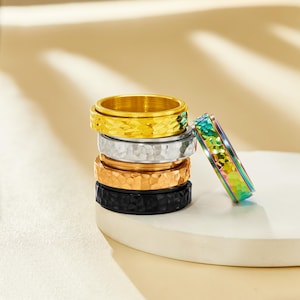 May include: A stack of five textured spinner rings in gold, silver, copper, and black. A rainbow-colored ring leans against the stack. The rings have a hammered metal finish and a rotating center band. Jewelry.