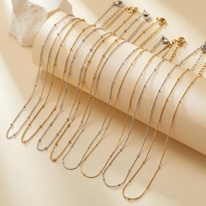 Puede incluir: Un conjunto de 12 delicados collares de cadena dorada y plateada con pequeñas cuentas. Los collares están dispuestos en forma de abanico sobre una superficie beige.
